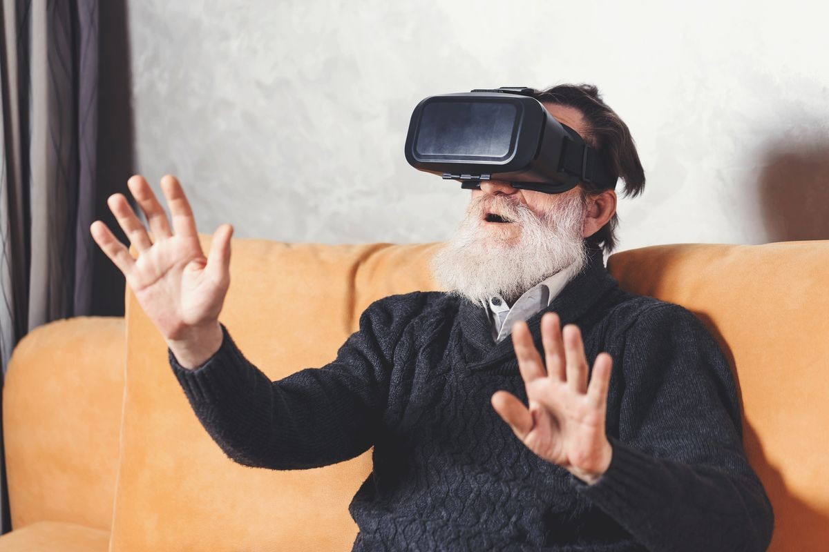 Surprised senior bearded man in grey pullover testing VR device while sitting on the yellow sofa in the light living room, future technology concept