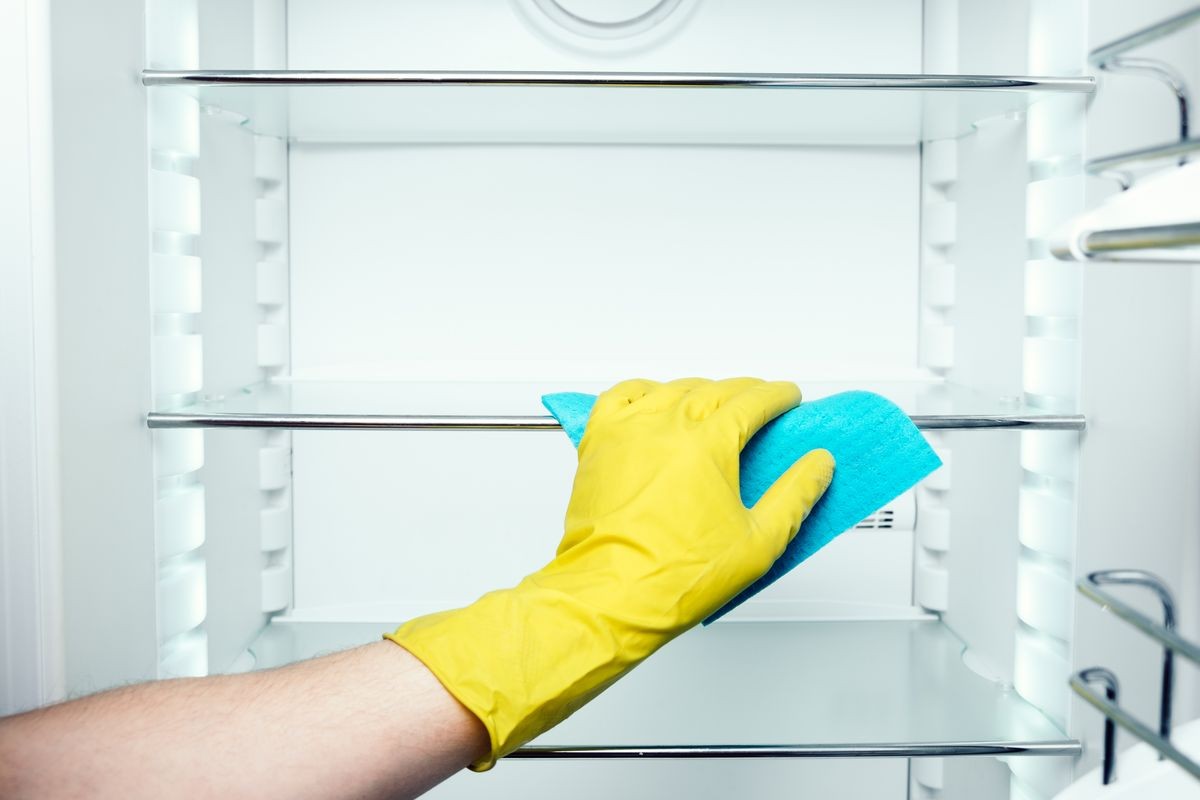Man's hand in yellow gloves washing refrigerator inside with blue duster